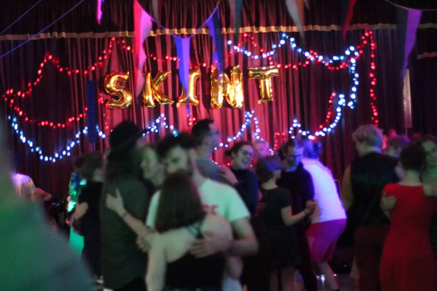 Couples dancing at a bal. There are fairy lights and banners, and large balloon letters spelling Skint